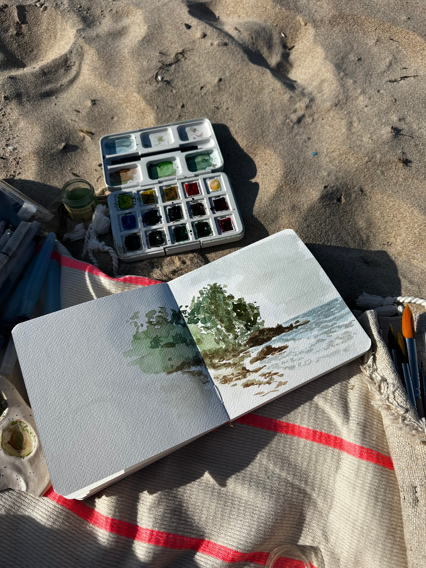 Sketchbook with driftwood study on a striped beach blanket with brushes
